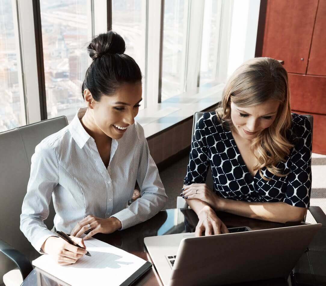 Two women sitting at a desk in an office, looking at a computer. One woman is writing on a notepad.